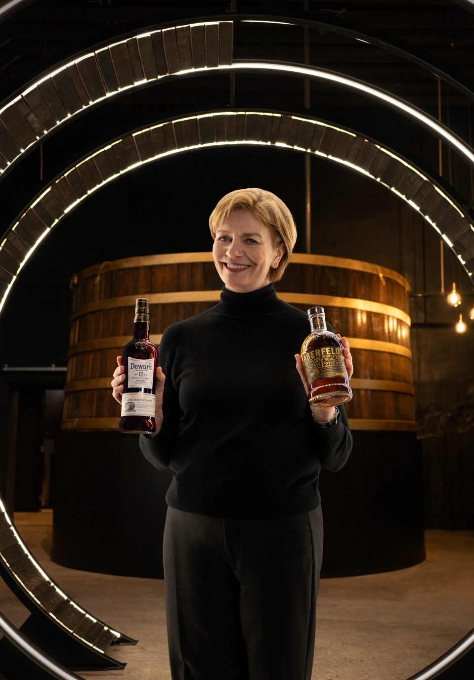 A photo of Stephanie Warehouse holding two bottles of Dewars. Dewar’s Aberfeldy Distillery